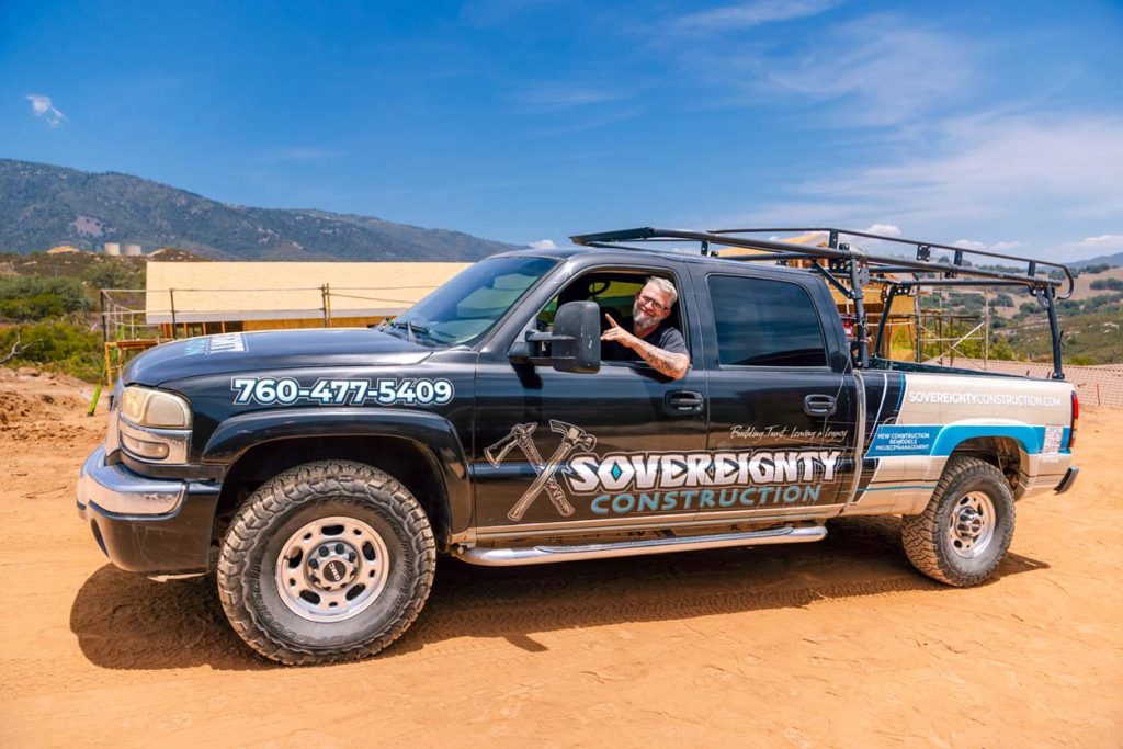 Sovereignty Construction branded pickup truck parked on a dirt job site with a team member waving from the driver's seat, contact number and logo visible on the door.