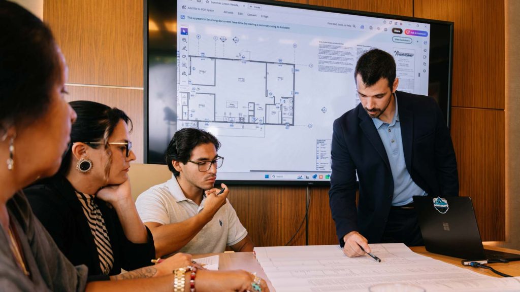 Sovereignty Construction representative pointing out details on a floor plan printout while clients review it, with the same layout displayed on a screen behind them.