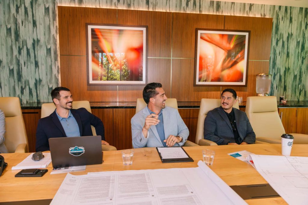 Three Sovereignty Construction team members in business attire sharing a laugh during a boardroom meeting, with a laptop, blueprints, and documents on the table, reflecting the firm's leadership in serving Tribal Nations.
